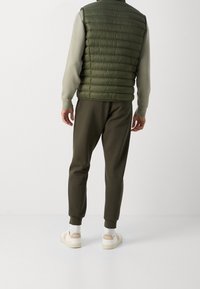 Man i olivgrön vadderad väst, ljusgrön långärmad skjorta, mörkgröna joggarbyxor, vita strumpor och beige-vita sneakers som står med ryggen mot kameran.