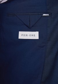 Tissu bleu marine au toucher lisse, avec une poche à rabat et une étiquette blanche indiquant "PIER•ONE" sur le côté.