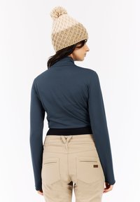 Vrouw die met haar rug naar de kijker staat, met een beige gebreide pomponmuts, een donkerblauw langemouwen shirt en beige broek, tegen een effen witte achtergrond.