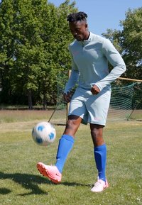 Light gray sportswear with pink accents, blue socks, and pink soccer shoes. The player controls a white soccer ball on a grassy field.
