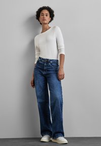 Blaue Denim-Hosen im Weitleg-Schnitt, hoch geschnitten, mit einer dezenten Waschung. Kombiniert mit einem weißen Langarm-Oberteil und weißen Sneakers vor einem neutralen Hintergrund.