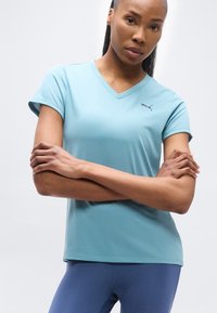 Γυναίκα με σταυρωμένα χέρια φοράει ανοιχτόχρωμη μπλε μπλούζα V-neck της Puma και σκούρο μπλε κολάν, μπροστά σε απλό λευκό φόντο.