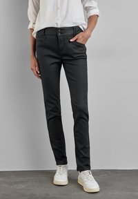 Personne portant un pantalon noir moulant, des baskets blanches et une chemise blanche à manches longues, avec une main dans la poche, sur un fond gris.