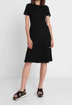 Kobieta ubrana w czarną sukienkę z krótkim rękawem do kolan z bocznymi rozcięciami i czarne sandały na platformie z białą podeszwą.