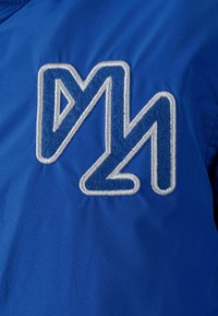 Mėlynos striukės audinys su išsiskiriančiu išsiuvinėtu logotipu, kuris turi kampuotas formas įvairių mėlynų atspalvių ir yra apibriestas baltu siūlu.