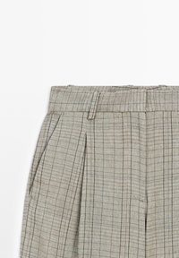 Pantalon beige taille haute à carreaux avec fines lignes noires verticales et horizontales, plis, poches latérales et passants visibles sur fond blanc.