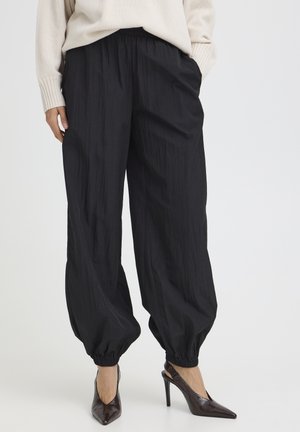 Osoba oblečená v voľných čiernych nohaviciach zviazaných pri členkoch, v kombinácii s tmavohnedými špicatými vysokými podpätkami a krémovým svetrom.