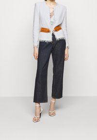 Biała teksturowana kurtka z postrzępionymi krawędziami, połączona z brązowym paskiem w talii. Ciemne, szerokie spodnie z denimu i białe, paskowe sandały na wysokim obcasie.