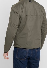 Olive zelená bomber bunda s hladkým povrchem, s detailem na zadním dílu a elastickými manžetami, v kombinaci s tmavě modrými džínami.
