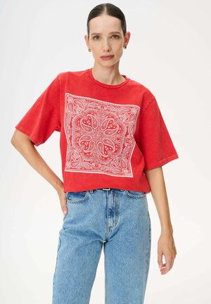 Kobieta z ciemnymi włosami związanymi z tyłu nosi czerwoną, oversize koszulkę z białym wzorem paisley oraz jasnoniebieskie, wysokie jeansy na białym tle.