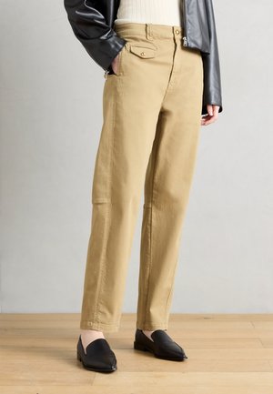 Személy beige egyenes szárú nadrágban, gombos zsebes légyelemmel, fekete bőrkabátban, krémszínű bordázott felsőben és fekete mokaszinban, fapadlón állva.