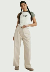 T-shirt bianco e verde oliva a maniche corte con logo grafico, abbinato a pantaloni beige plissettati. Modello che indossa pantofole grigio chiaro.