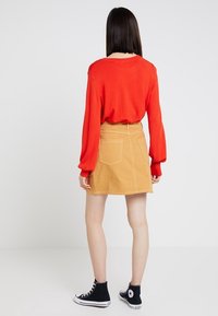 Pull en tricot rouge à manches longues, associé à une jupe en denim beige avec deux poches arrière et des coutures contrastées, porté avec des baskets noires.