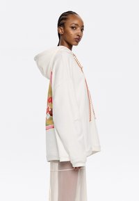 Sudadera blanca con un gran estampado gráfico en la manga izquierda, que presenta elementos en rosa, dorado y florales. Tiene capucha con cordones.