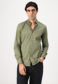 Zöld ing, kis csillagmintával, hosszú ujjakkal és slim fit szabással, sötét farmernadrággal párosítva. Az ingnek standard gallérja van és barna gombokkal rendelkezik.