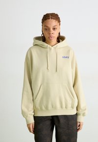 Hoodie beige claro com um bolso frontal e capuz com cordão. Apresenta texto azul "obey" no lado esquerdo do peito. Tecido suave, ajuste descontraído.