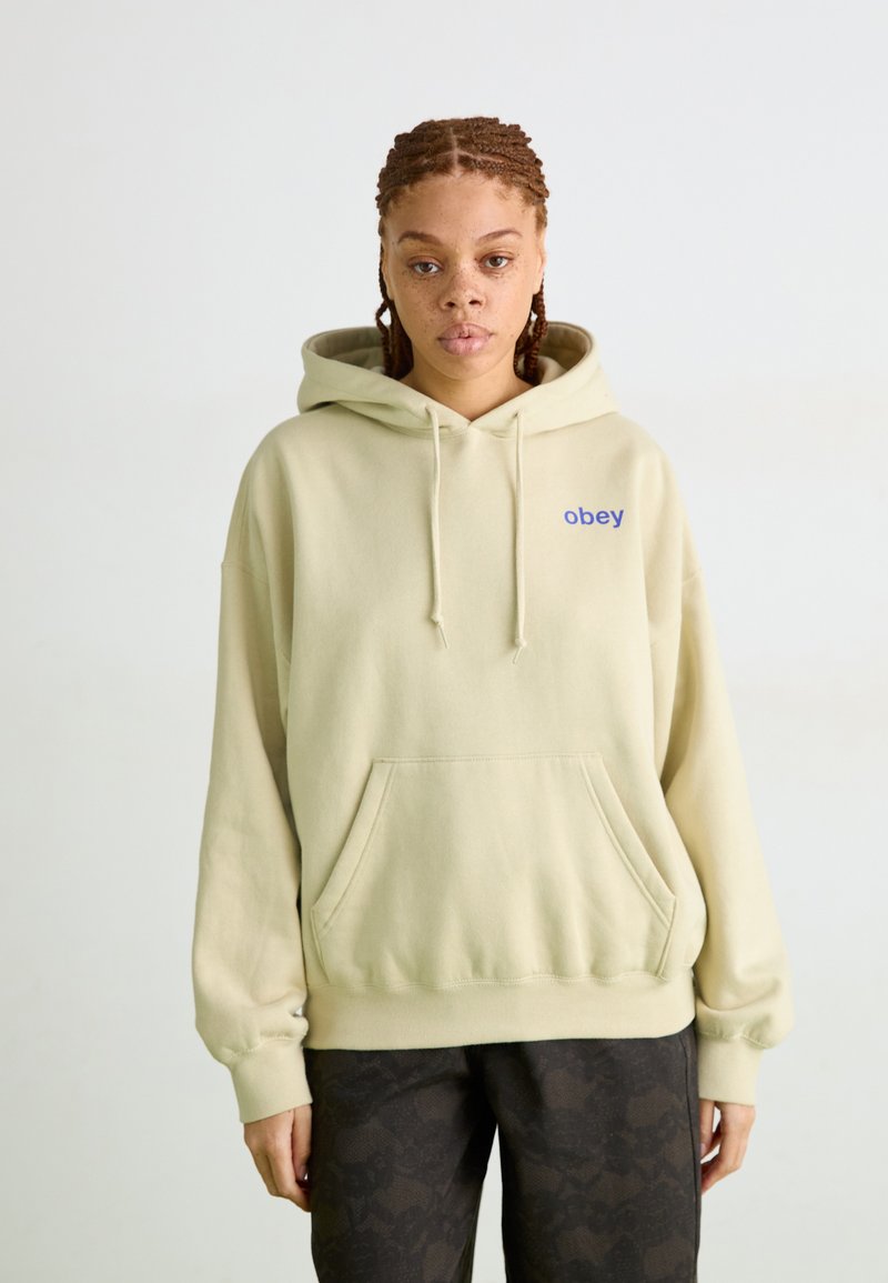 Hoodie beige claro com um bolso frontal e capuz com cordão. Apresenta texto azul "obey" no lado esquerdo do peito. Tecido suave, ajuste descontraído.