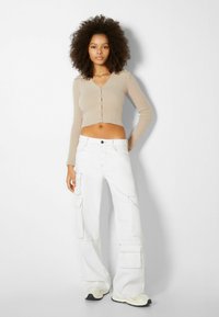 Beige kort kofta med långa ärmar och v-ringning, kombinerad med vita vida cargobyxor med flera fickor och en avslappnad passform.