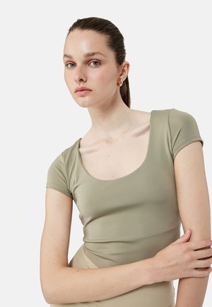 Junge Frau mit heller Haut und blauen Augen, die goldene Creolen und ein eng anliegendes, kurzärmeliges Oberteil in Salbeigrün trägt, posiert vor weißem Hintergrund.