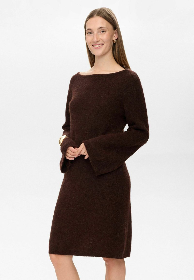 Rochie tricotată maro, cu decolteu tip barcă, mâneci lungi tip clopot și o siluetă ajustată. Textură moale, cu un design până la genunchi. Fără modele sau accesorii vizibile.
