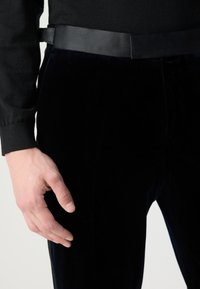 Juodi aksominiai kelnės, turinčios lygų paviršių, derinamos su elegantišku juodu diržu ir prigludusia juoda ilgomis rankovėmis marškiniais.