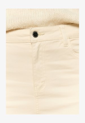 Beige puuvillaiset farkut, joissa on sileä pinta, musta nappikiinnitys, ommellut taskut ja näkyvät saumat.