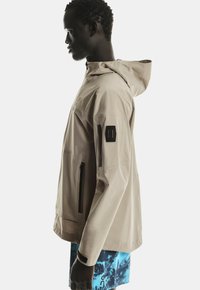 Beige waterdichte jack met een capuchon, voorzien van een ritszak op de borst en zwarte accenten, gedragen over blauwe gestreepte shorts.