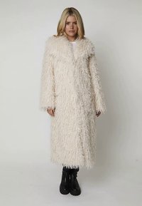 Manteau long et shaggy de couleur blanc cassé, avec un large col et une ouverture devant. La texture est douce et duveteuse, associé à des bottes à plateforme noires.