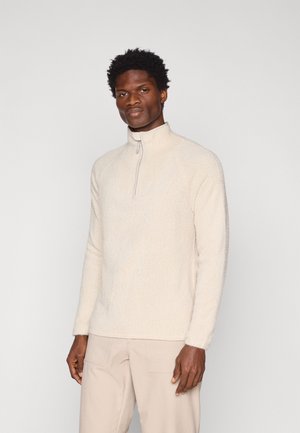Beige fleece-tröja med dragkedja i kragen, texturerad yta, åtsittande ärmar och avslappnad passform. Mjuk, lättviktsmaterial med ett subtilt mönster.