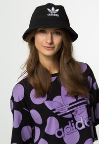 Cappello a secchiello nero con logo adidas bianco, indossato con una maglietta nera caratterizzata da grandi cerchi viola e marchio sotto la scollatura.