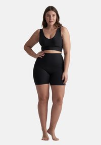 Donna in piedi a piedi nudi, con indosso un reggiseno sportivo nero e pantaloni biker neri a vita alta, su uno sfondo bianco uniforme.