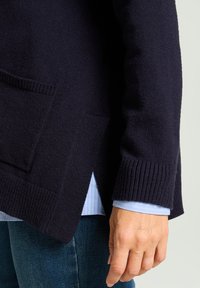 Marineblauer Strickcardigan mit gerippten Bündchen und Saum, ausgestattet mit zwei Vorderentaschen und seitlichen Schlitzen für mehr Bewegungsfreiheit. Darüber getragen wird ein hellblaues Hemd.