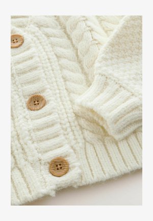 Kermanvärinen neulecardigan, jossa on paksu kabelineulekuvio, ribattu hihansuut ja puunapit kiinnitystä varten. Pehmeä, teksturoitu kangas.