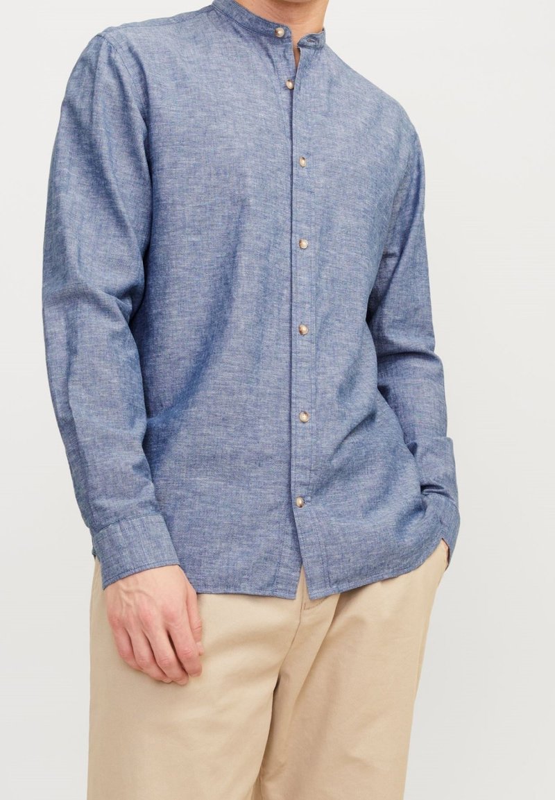Homme portant une chemise bleue à manches longues avec col mandarin et boutons marron ainsi qu’un pantalon beige, main reposant sur le côté et dans la poche.