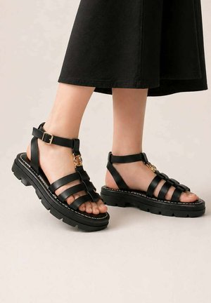 Pieds portant des sandales en cuir noir à lanières avec des boucles dorées et des semelles épaisses texturées, associées à un pantalon court noir.