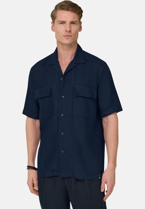 Mand iført en navyblå kortærmet skjorte med knapper og to brystlommer samt mørke bukser mod en ensartet lys baggrund.