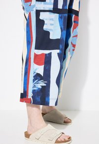 Pantaloni larghi con un motivo astratto multicolore in blu, rosso e crema. Abbinati a sandali grigi chiari con due cinturini.