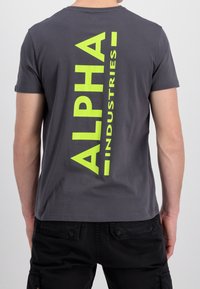 Šedé tričko s krátkým rukávem, které na zádech zdobí velké vertikální logo "ALPHA INDUSTRIES" v žluté barvě. Vyrobeno z měkké bavlny.