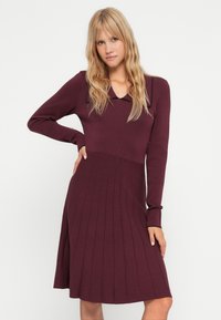 Abito lavorato a maglia bordeaux con maniche lunghe, collo a camicia e gonna plissettata. Tessuto liscio con dettagli a costine sottili sulle maniche.
