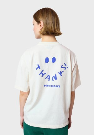 Cream-colored oversized t-shirt with short sleeves featuring blue "THANKS!" text and smiley face design on the back. Soft cotton material.