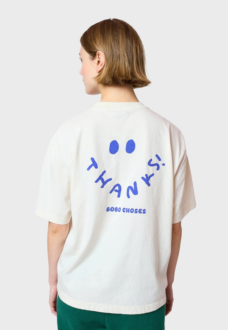 Camiseta de gran tamaño color crema con mangas cortas, que presenta un texto azul de "¡GRACIAS!" y un diseño de cara sonriente en la parte posterior. Material de algodón suave.