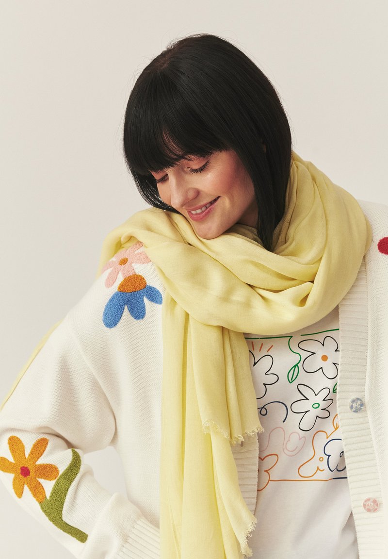 Femme aux cheveux noirs sourit en regardant vers le bas, portant un pull blanc à motifs floraux, une écharpe jaune clair et une chemise assortie avec des motifs floraux colorés.