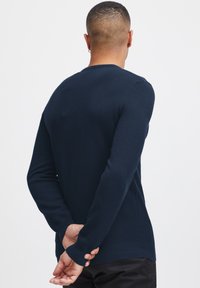 Navy blue sweater with textured knit pattern, crew neckline, long sleeves, and ribbed cuffs and hem. Back view, hands clasped behind.