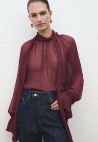 Bluză transparentă bordeaux cu guler înalt și mâneci lungi, asociată cu blugi din denim închis la culoare, cu nasture frontal și buzunare.