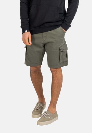 Mann trägt olivgrüne Cargoshorts, beige Sneaker und ein schwarzes Langarmshirt, steht mit einer Hand in der Tasche.