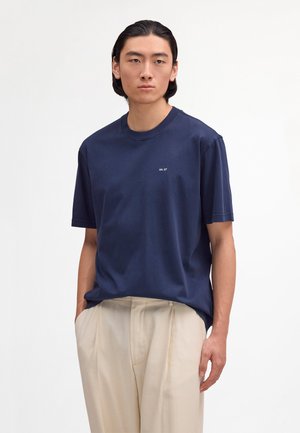 Giovane uomo con capelli scuri indossa una maglietta blu navy e pantaloni beige, in piedi con le mani nelle tasche contro uno sfondo semplice.