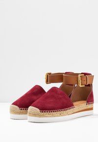 Espadrilles en suède rouge avec un bout rond, une bride de cheville marron, une boucle dorée et des détails en jute tressé autour de la semelle. Semelle en caoutchouc blanche.