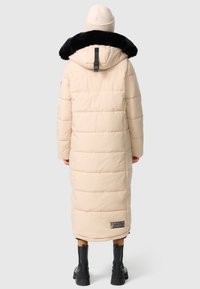 Beige langer Daunenmantel mit einem schwarzen Kunstfellkragen, gestepptem Material und einem Etikett am Saum. Getragen mit einer Mütze und schwarzen Ankle Boots.