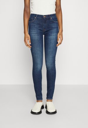 Jeans skinny bleu foncé en denim, dotés de cinq poches, d'une fermeture à bouton et à glissière, et d'une silhouette ajustée. Portés avec des chaussures blanches à enfiler.