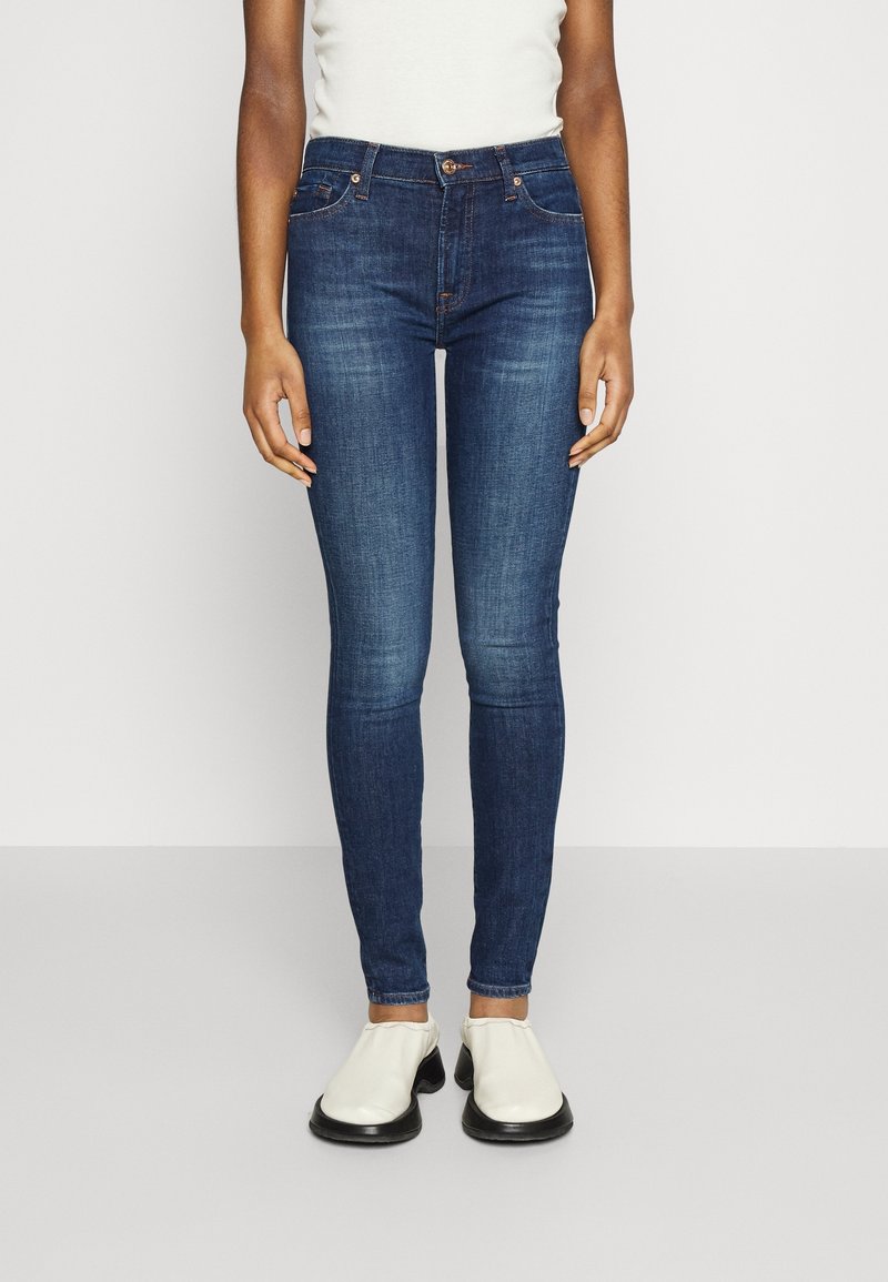 Jeans ajustados de mezclilla azul oscuro, con cinco bolsillos, cierre de botón y cremallera, y una silueta ceñida. Llevados con zapatillas blancas de tipo slip-on.